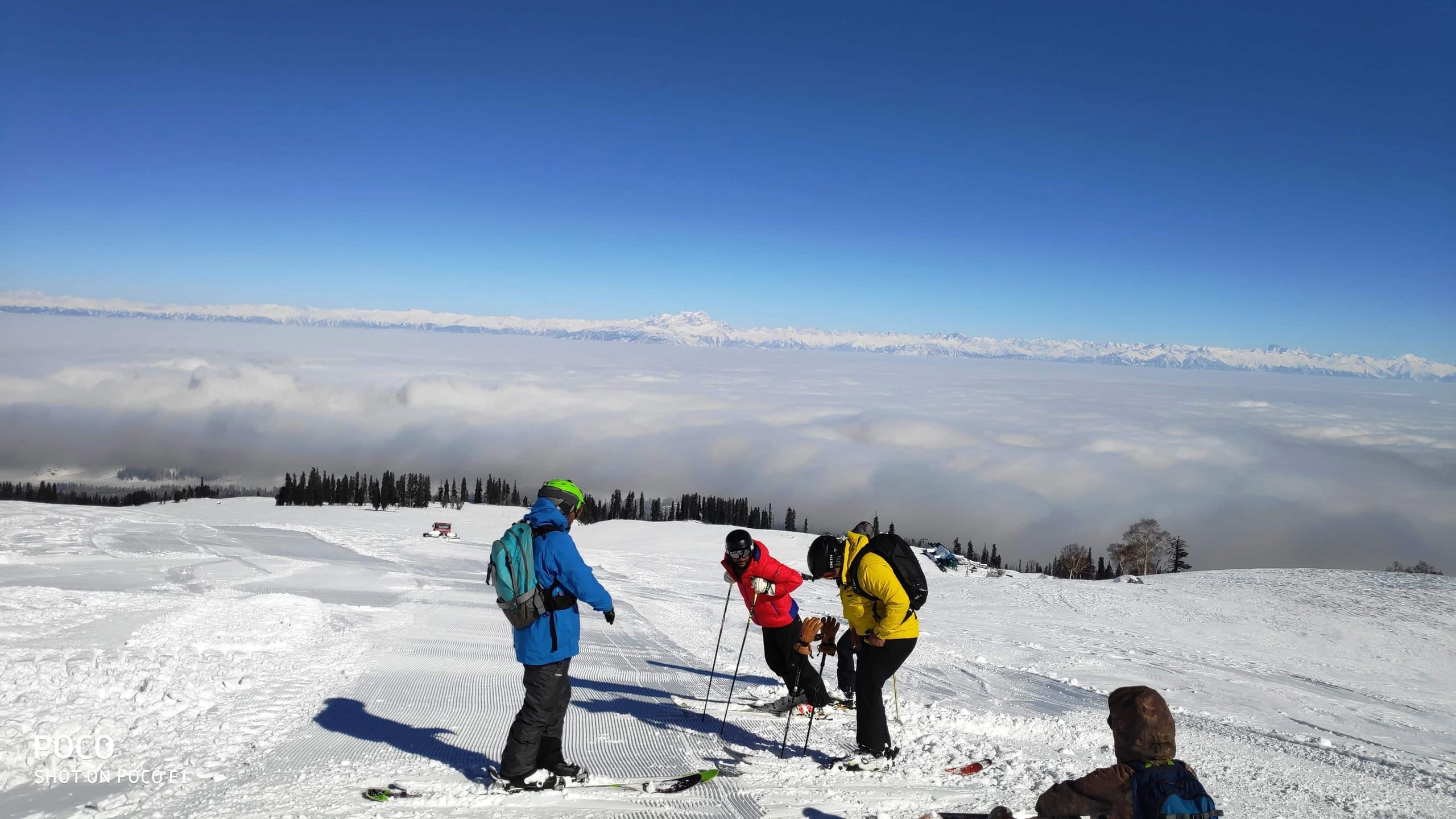 Three skiers standing on a snow ridge above a sea of cloud, looking out at the range beyond.