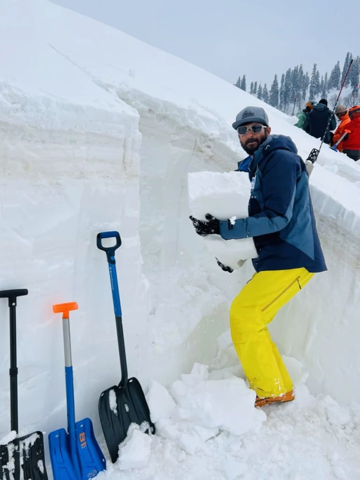 A guide standing in a dug snowpit holding a cut block of snow, with probes and shovels planted in the wall beside them.