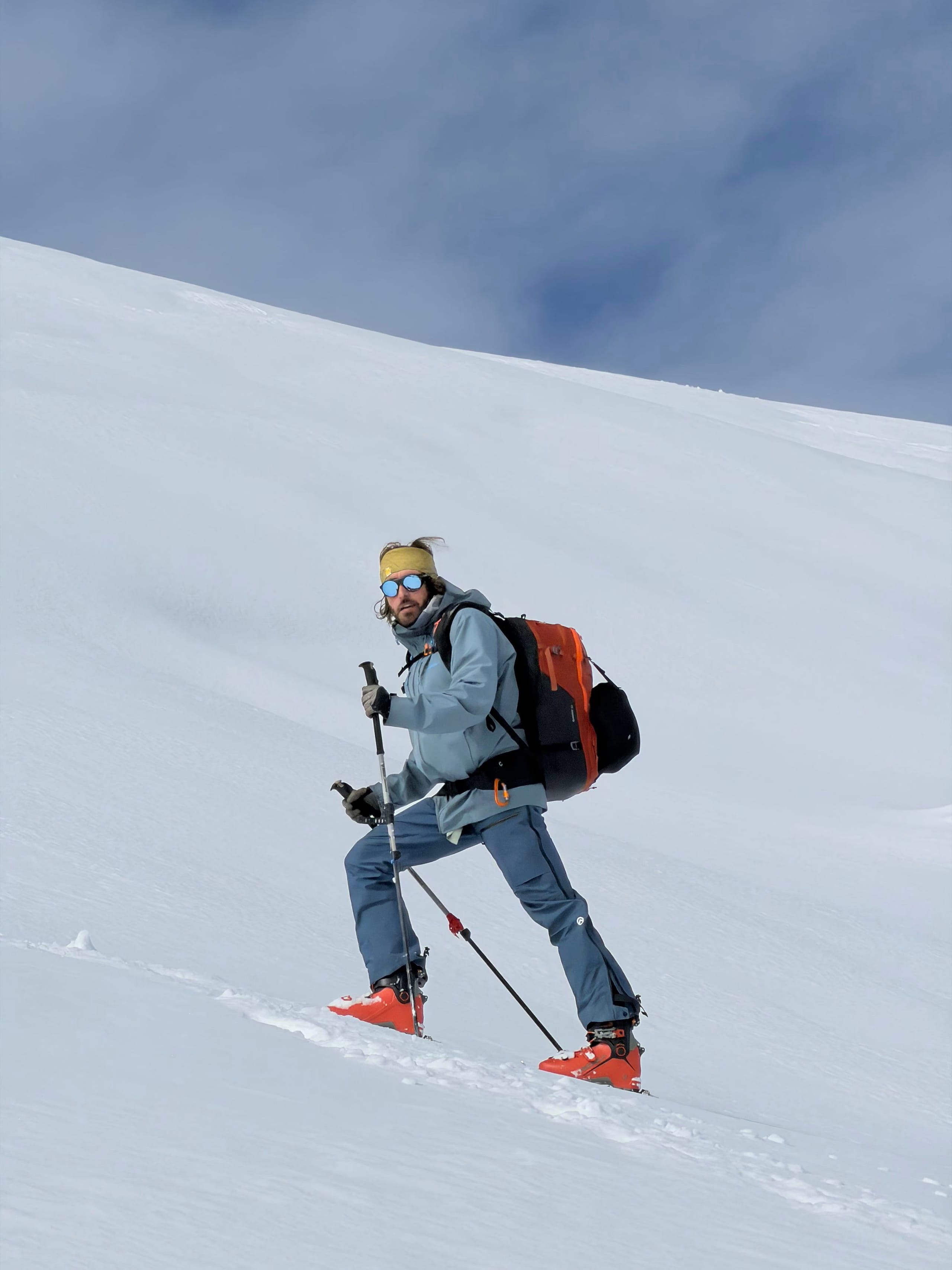 A skier with an orange pack climbing a steep untracked snow slope, kicking steps into the surface.
