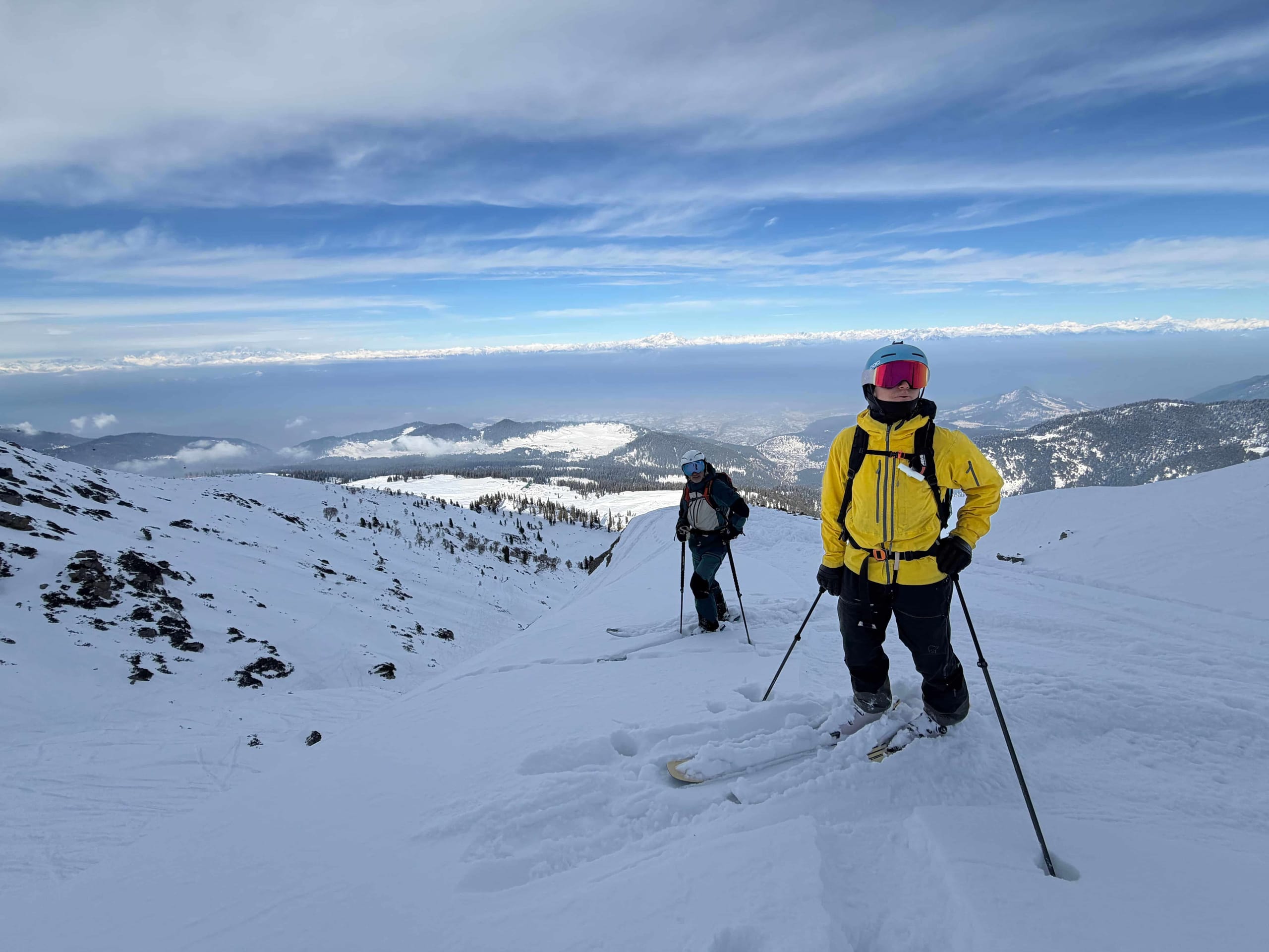 Two skiers standing on an open snowfield high on the mountain, looking out over ridgelines that run to the horizon.
