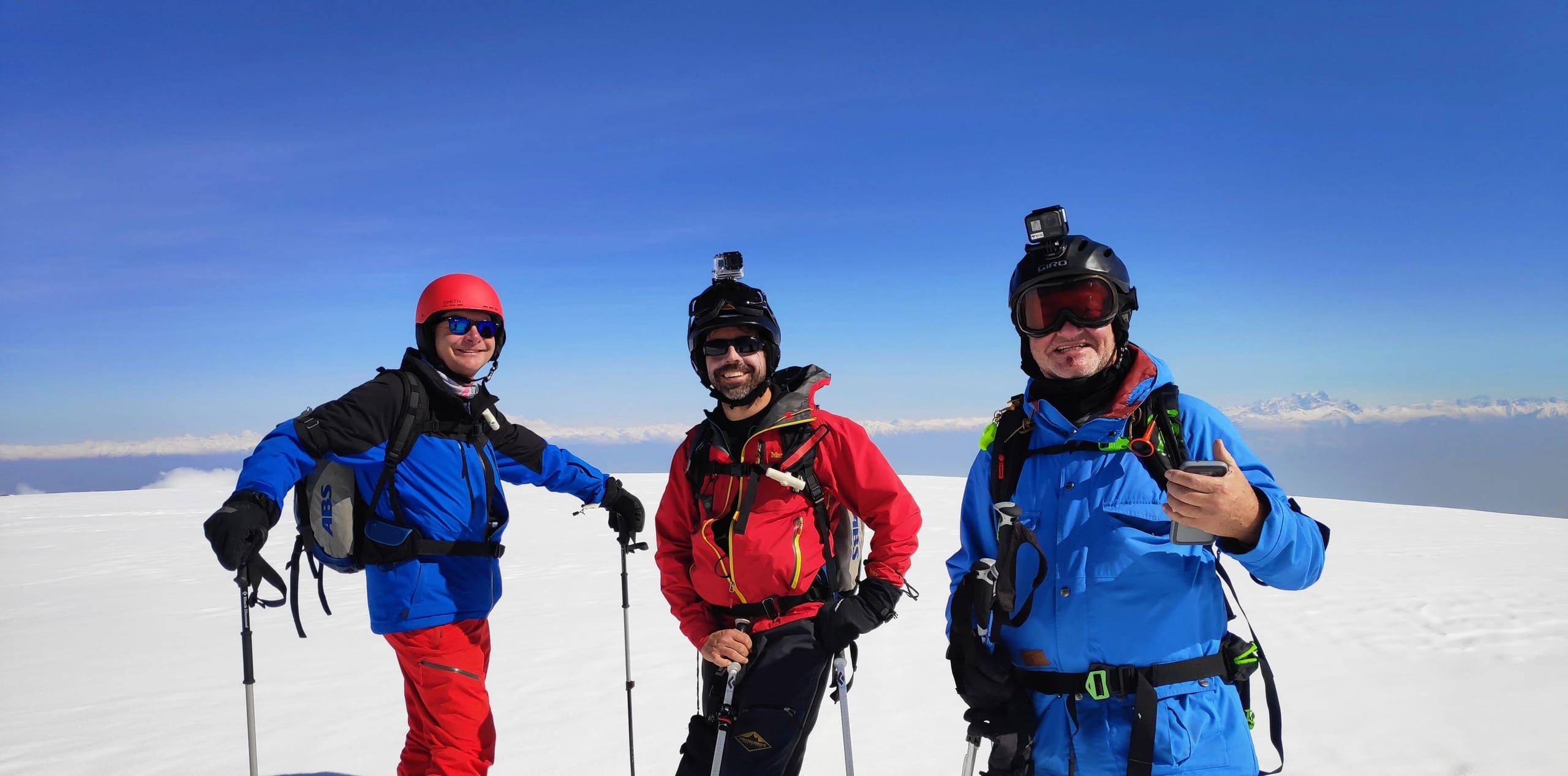 Three skiers wearing helmet cameras and packs standing on a snowfield above a sea of cloud.