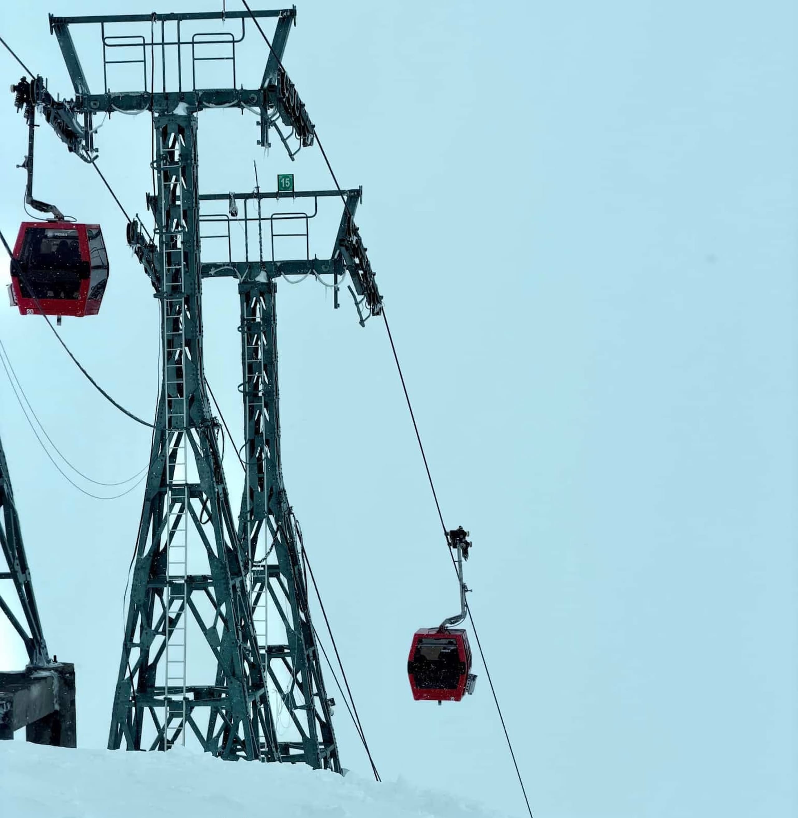 Two red Gulmarg Gondola cabins on the cable beside a steel lattice tower numbered 15, against a pale overcast sky with snow below.