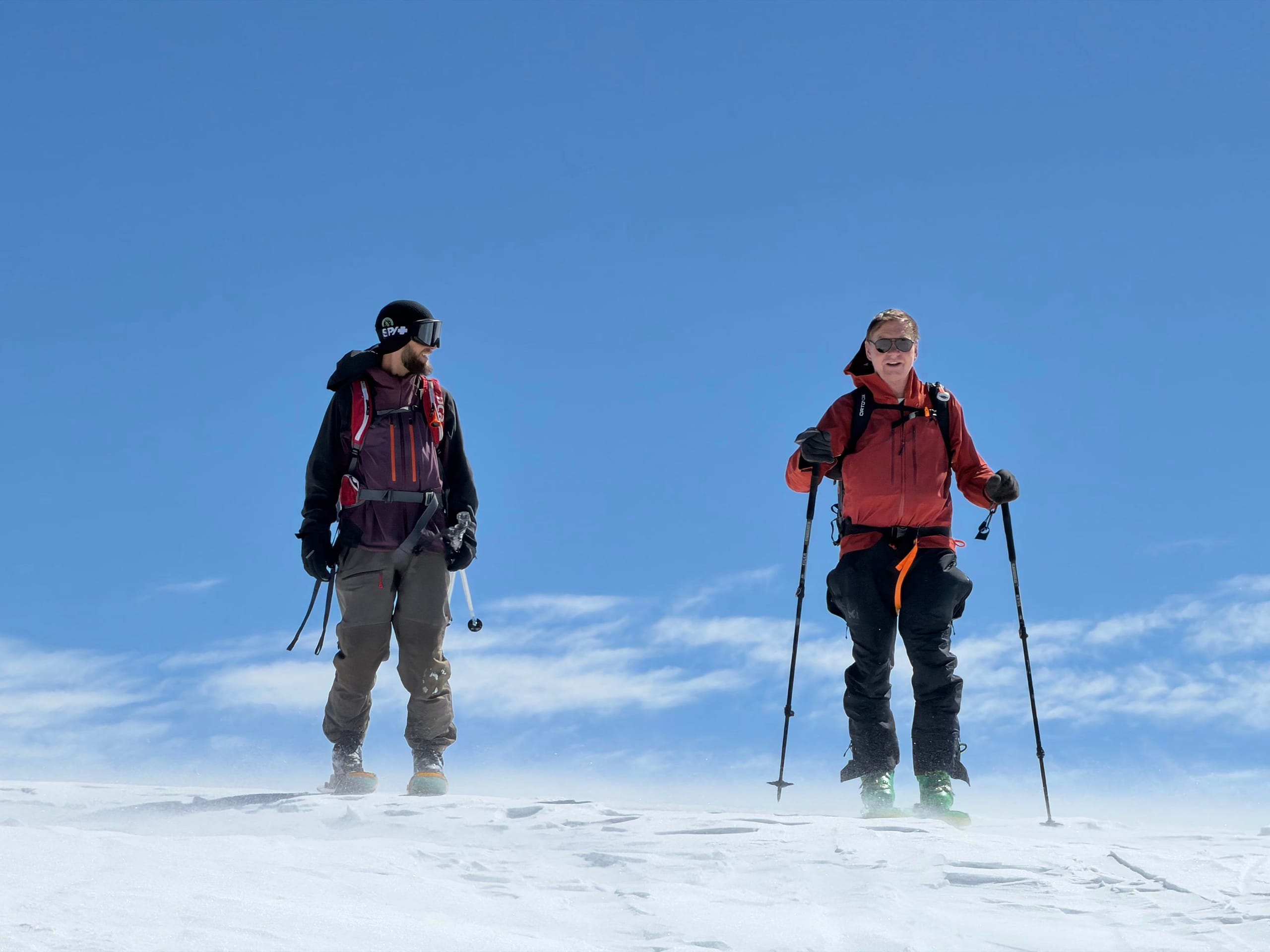 Two skiers wearing avalanche packs standing on a wind scoured snow ridge under a deep blue sky, spindrift blowing across the surface.