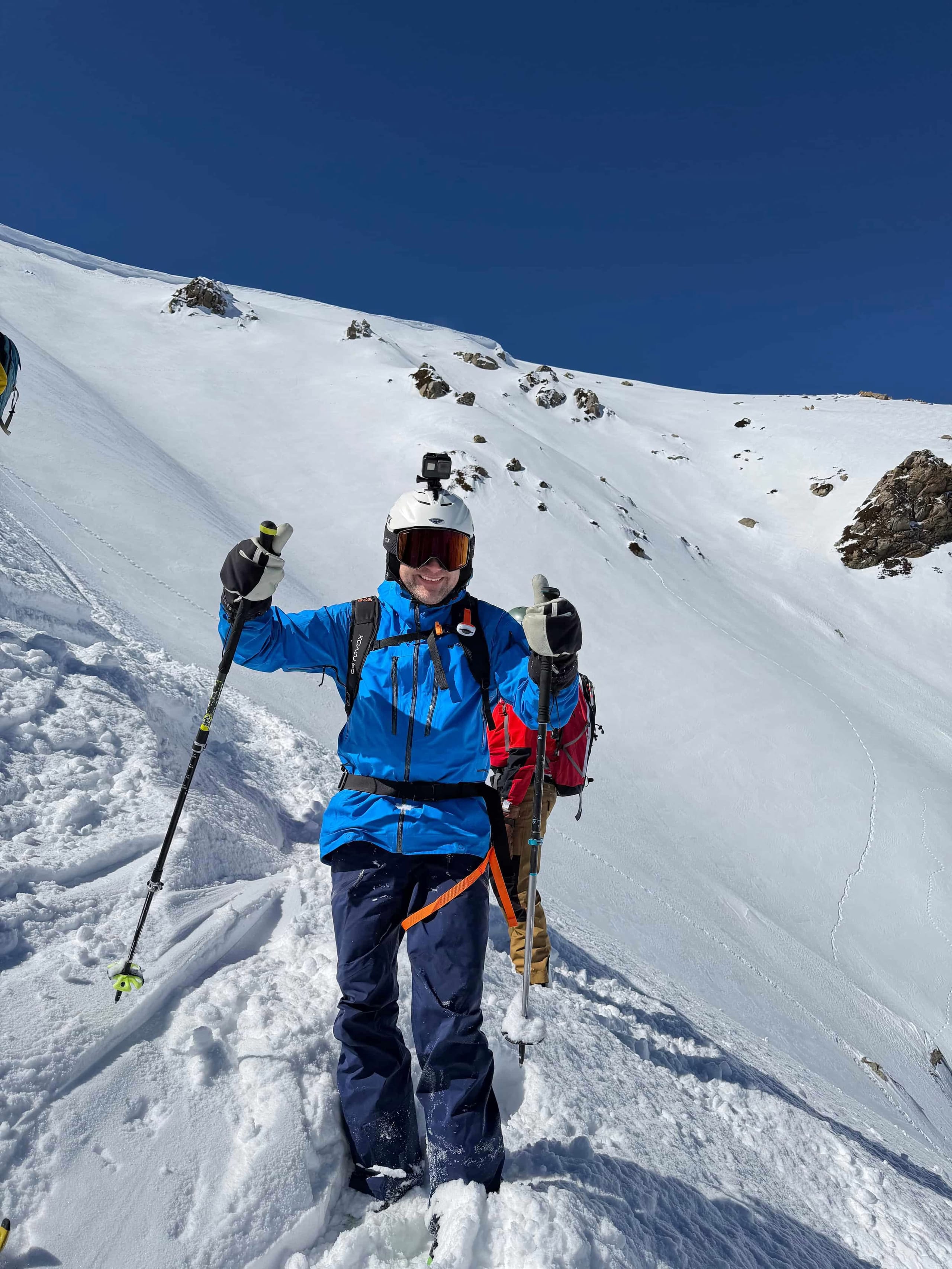 A skier in a blue jacket giving a thumbs up on a steep untracked off-piste slope, with a second skier in red higher up the face.