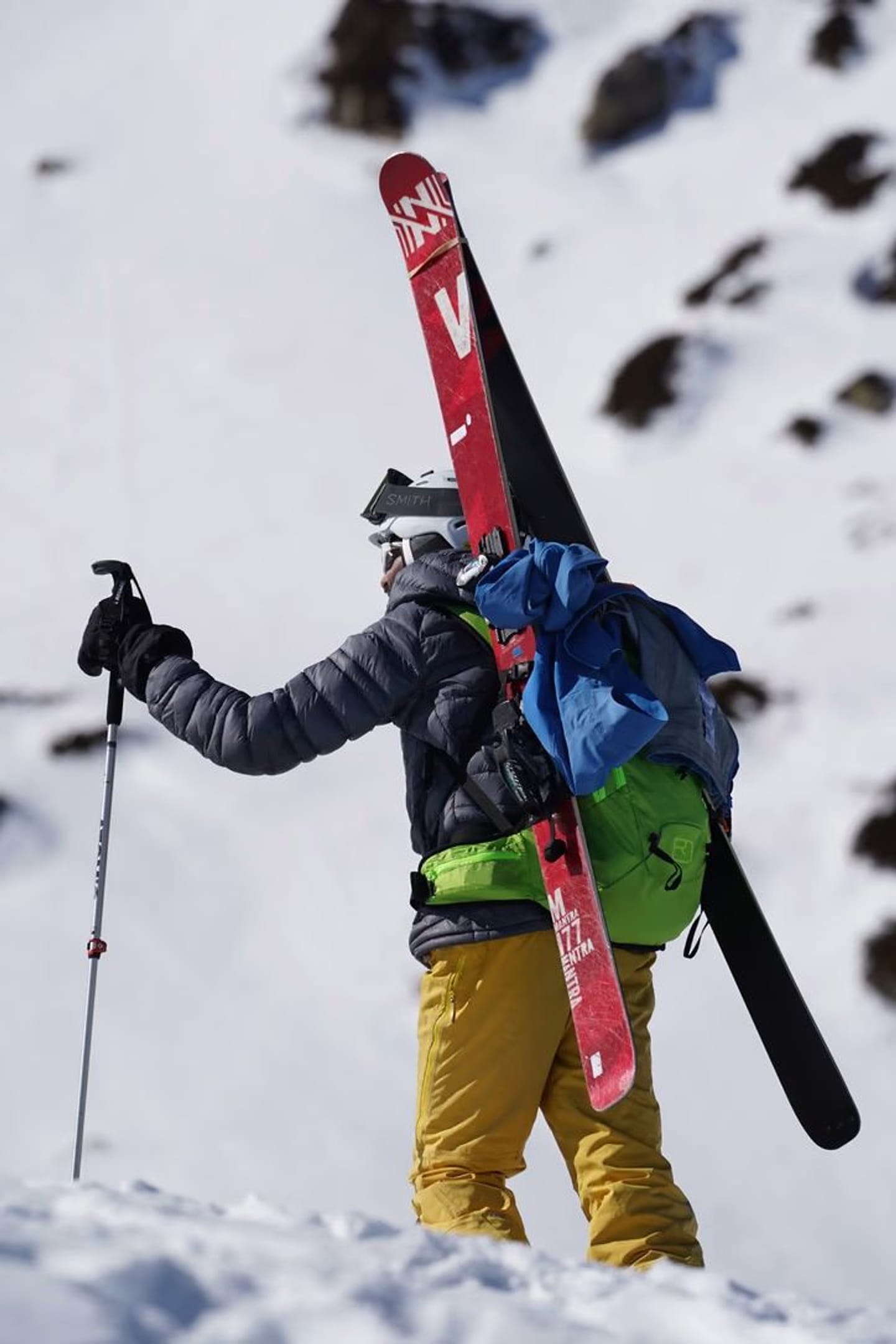 A skier bootpacking uphill with their skis strapped to a green backpack, holding a pole for balance.