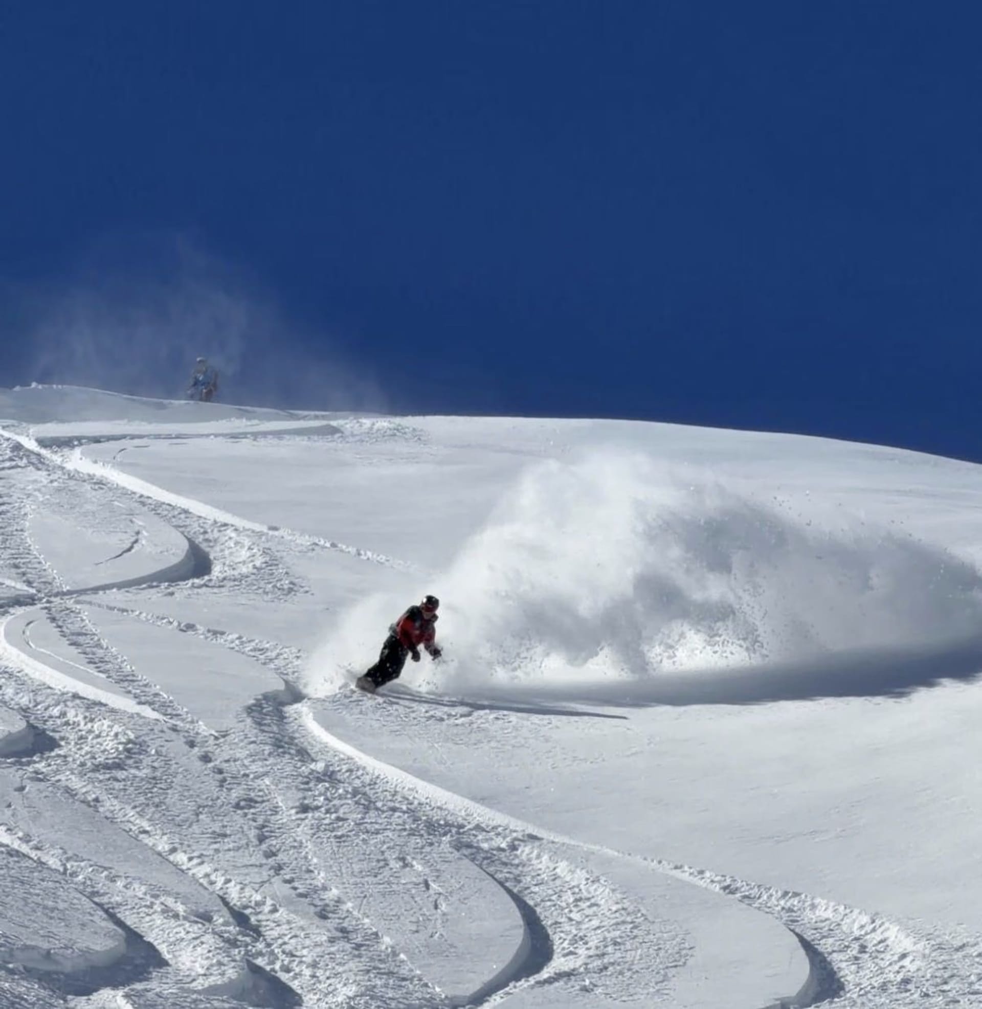 A snowboarder carving hard down an open powder face, throwing a large spray of snow with their track curving behind.