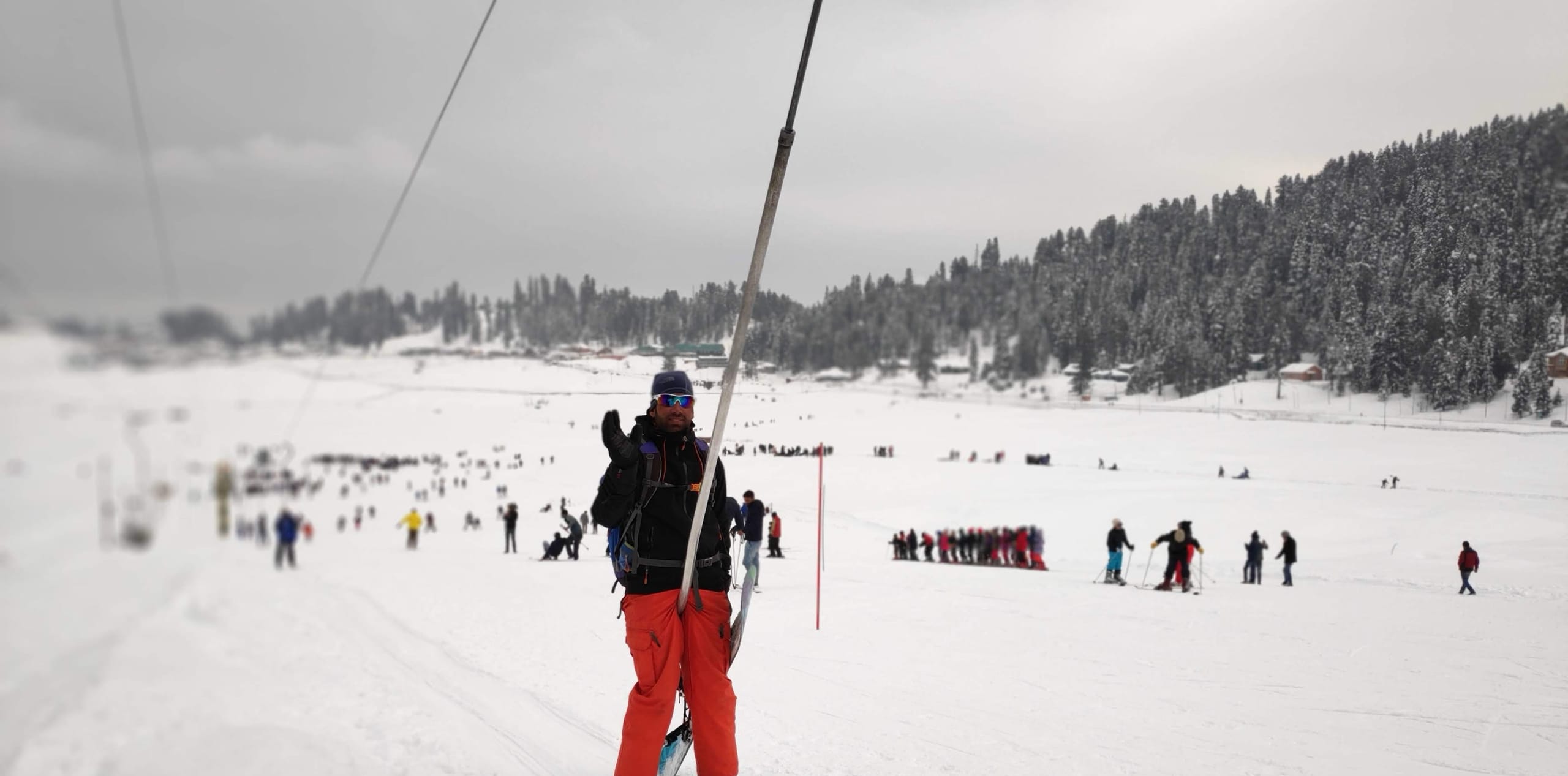 A skier in red trousers beside the rope tow on the Gulmarg nursery slope, with other skiers and pine forest in the background.