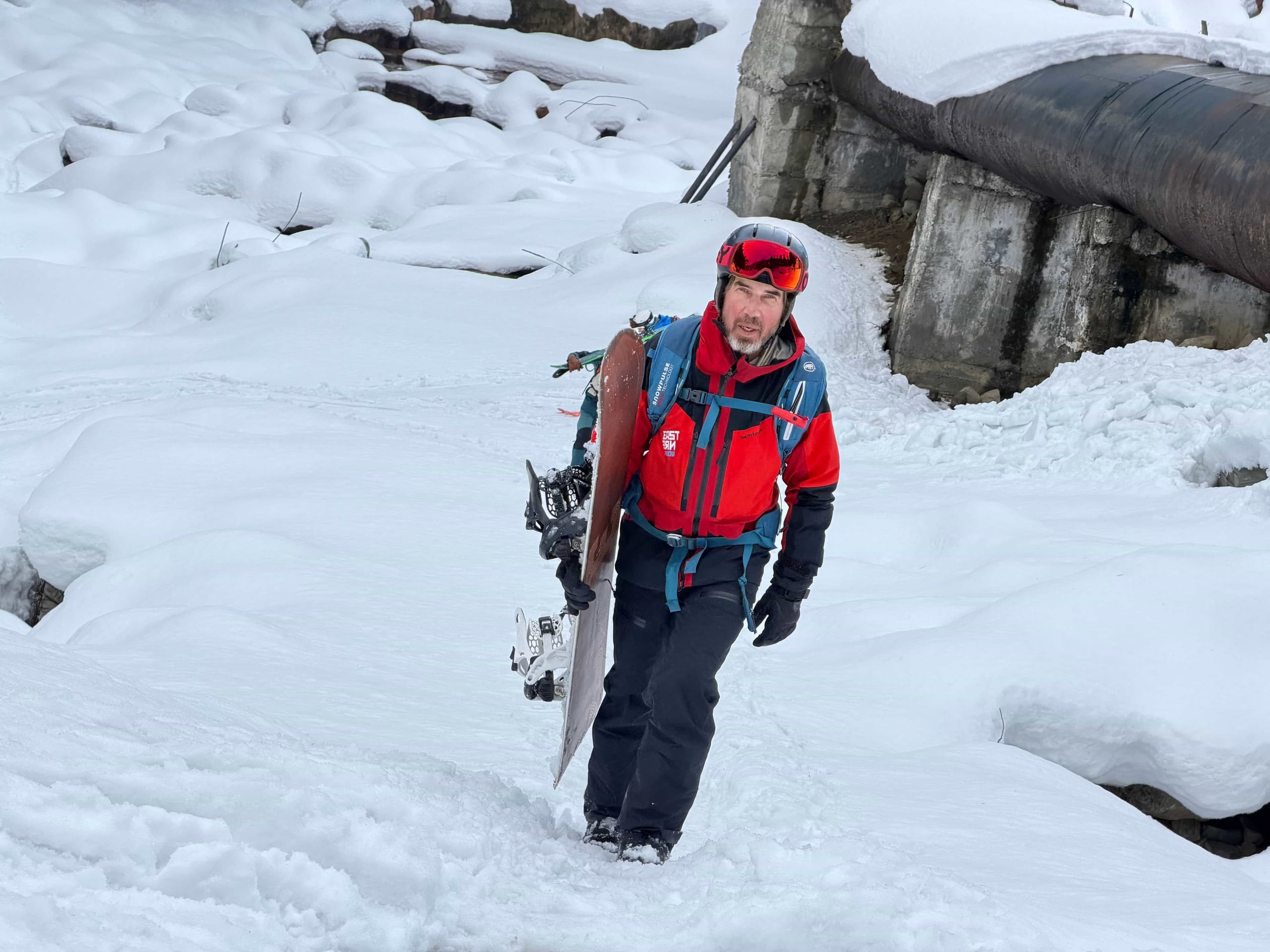A splitboarder walking uphill through deep snow with their board strapped to their pack, passing a rock outcrop.