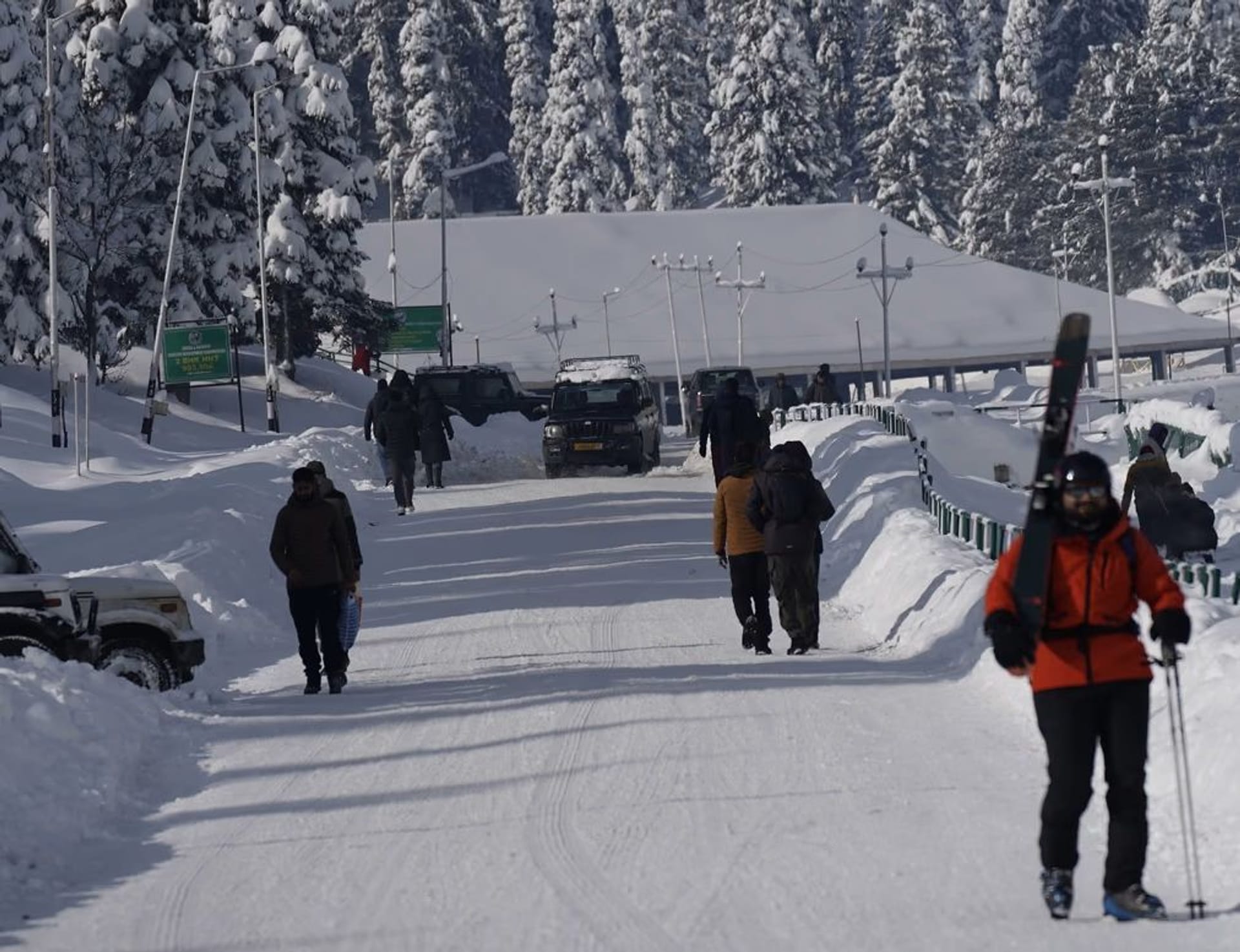 A snow cleared road through Gulmarg village lined with hotels and parked cars, with people walking past carrying skis.